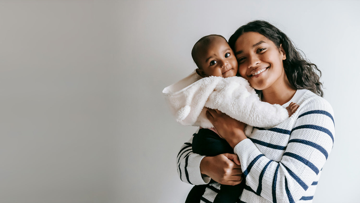 Smiling mum wearing a white and blue stripped jumper, cuddling a young baby both looking directly at the camera, stood against a white and grey background.