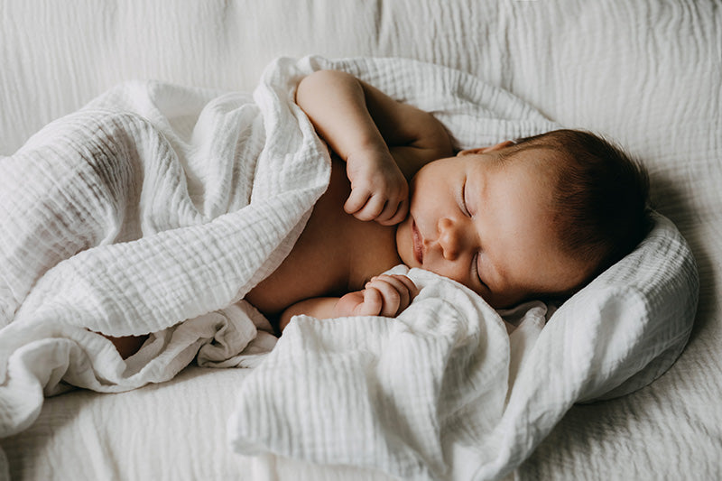 Sleeping newborn baby laying on soft, white linen sheets and wrapped up in a white muslin blanket.