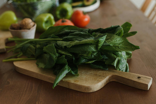 Wooden chopping board with fresh leafy greens on. Red tomatoes and green peppers in the background. 