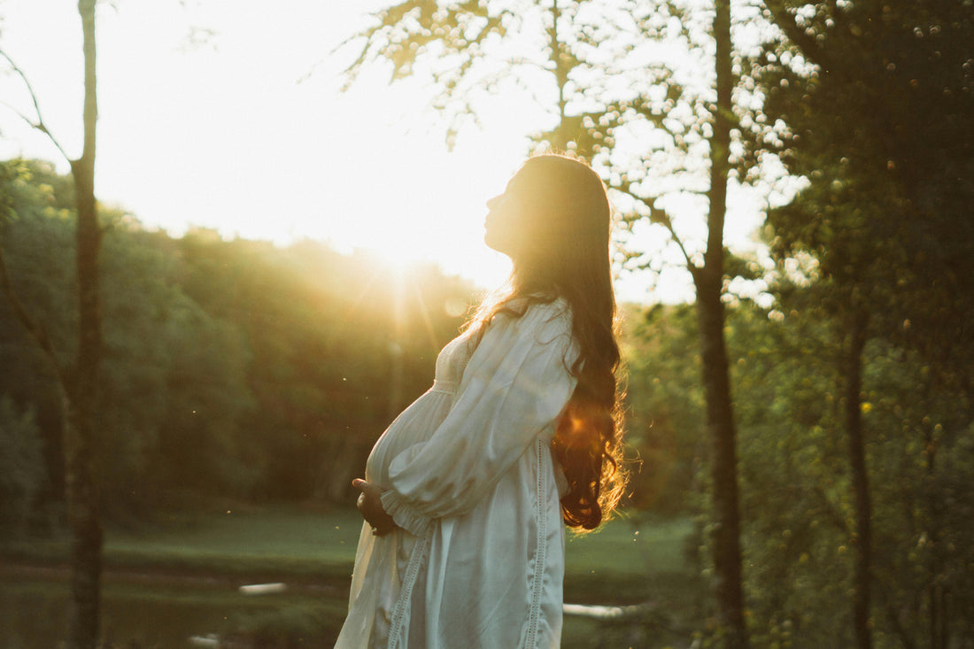 Pregnant lady wearing a white lace dress stood sideways, cradling her belly in amongst trees with sun shining from behind her. 