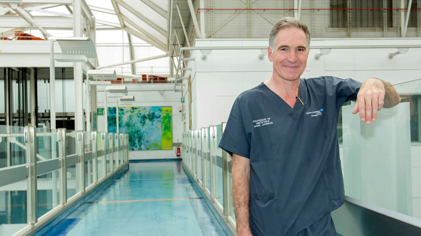 Professor Mark Johnson in a hospital setting, wearing NHS scrubs uniform, leaning against a glass barrier. 