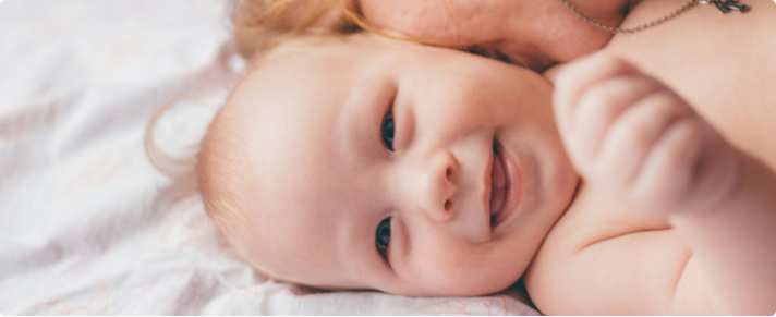 Baby laying on a white bedding being kissed on their neck by an adult. 