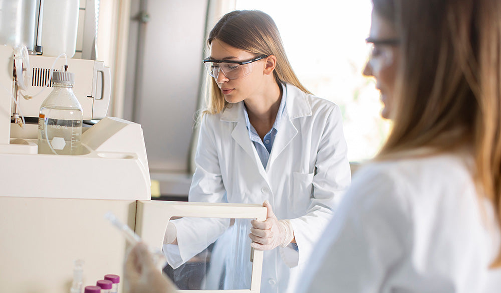 Scientists working in a lab, one holding a test tube, one wearing goggles opening a machine door. 