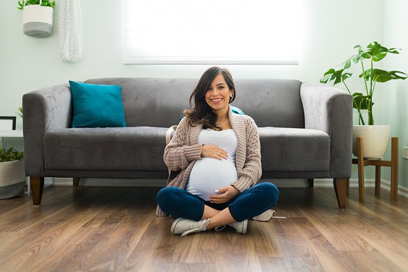 Pregnant lady sat on the floor smiling, cradling her bump leaning against a grey sofa in a warm living room. 