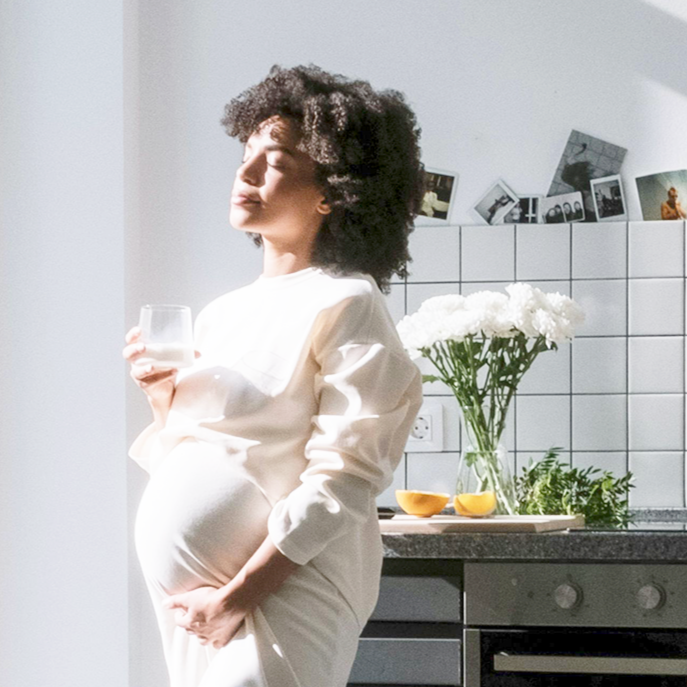 Pregnant lady holding a glass standing in a kitchen looking towards a window flooded with light with her eyes closed, holding her bump. 