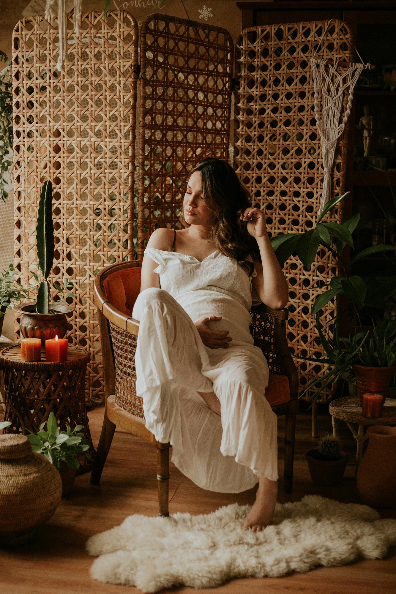Pregnant woman in a relaxed pose, sitting in a rattan chair and wearing a white off-shoulder dress. She is gently cradling her belly in a warm, natural setting with woven rattan panels, potted plants, and lit candles, creating a serene and cozy atmosphere.