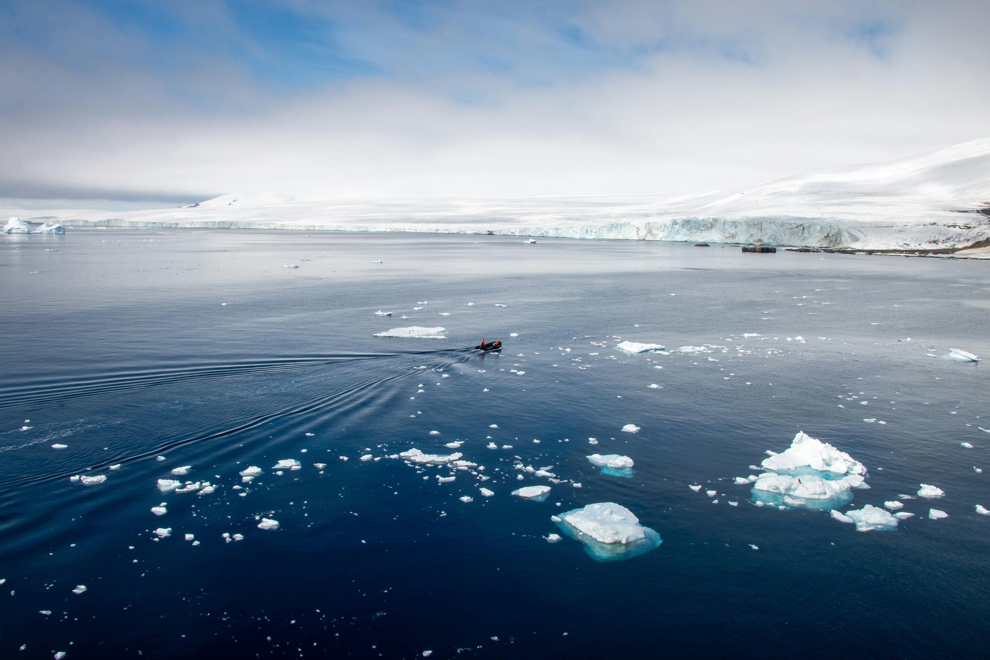 Vast blue ocean with small boat containing two people and white mountains in the background. 