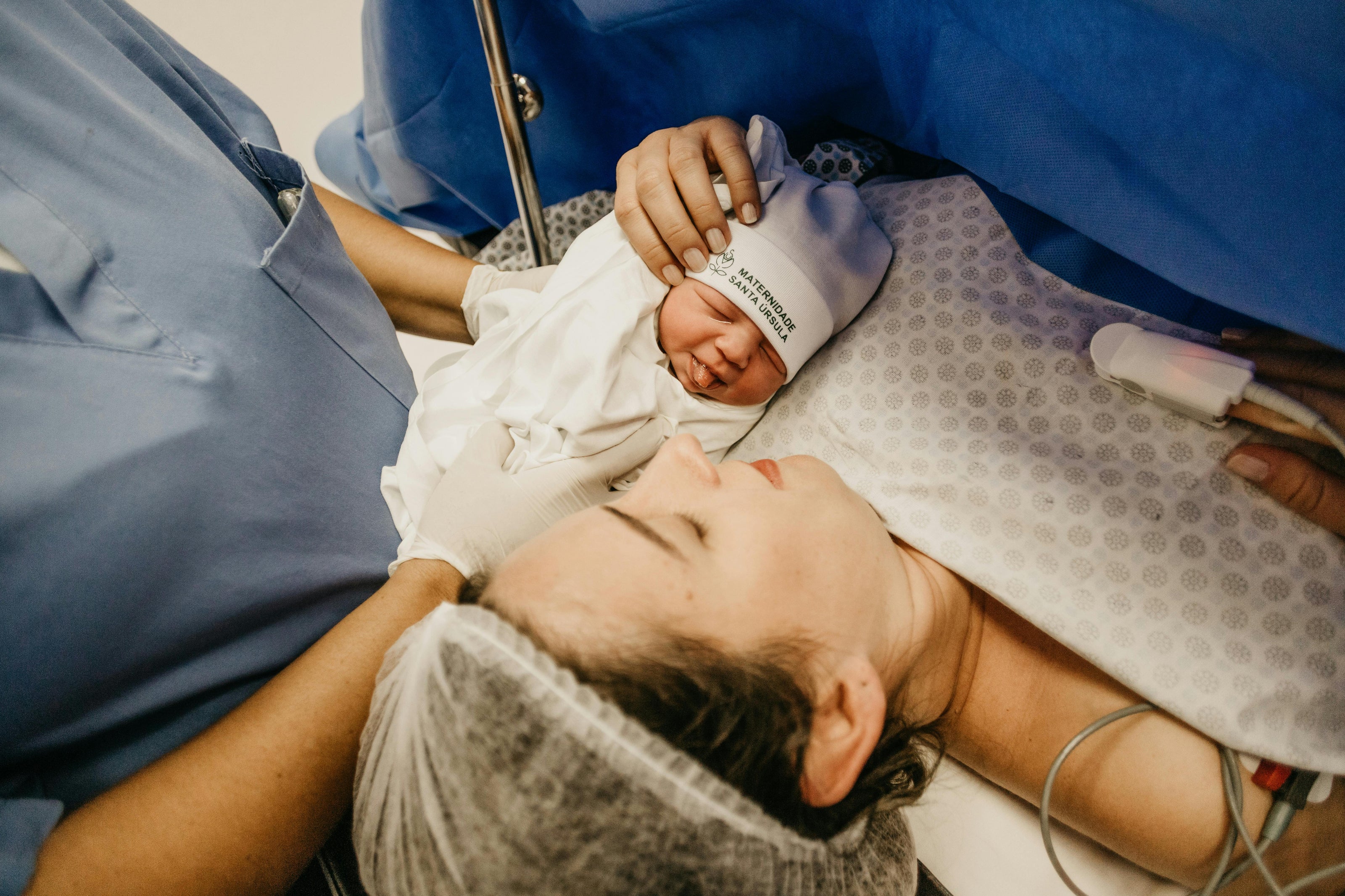 Doctor holding a newborn baby on a mum who is laying on a hospital bed with a hair net, oxygen monitor and blue curtain screen covering from her neck down. 