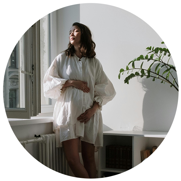 Pregnant lady in a floaty white dress, leaning against a radiator and windowsill looking towards the open window with her eyes closed. 